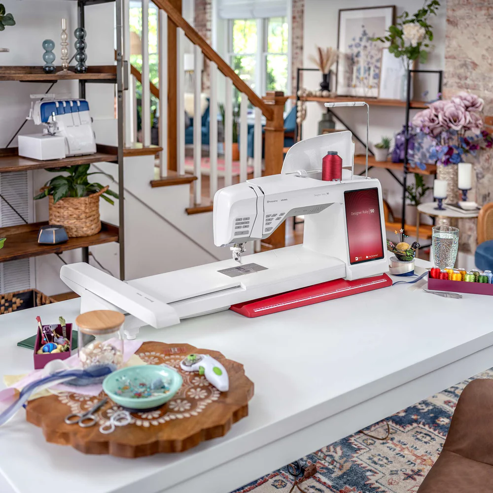 A modern sewing machine sits on a white table in a well-lit, organized craft room with shelves, sewing supplies, and decorative items.