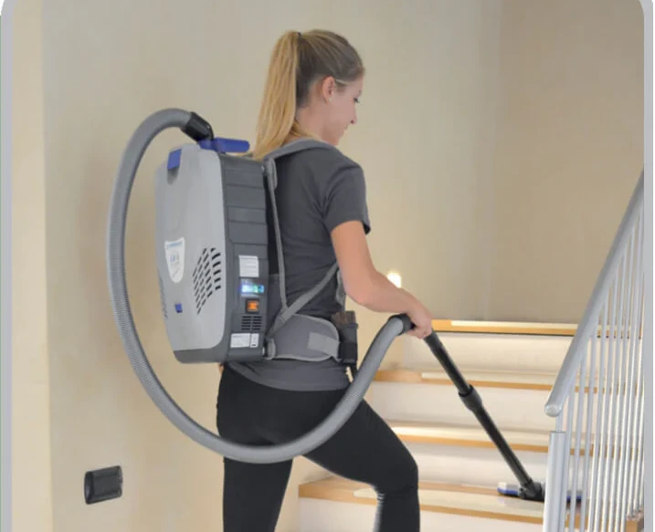 A woman uses a backpack vacuum cleaner to clean a set of indoor stairs, showcasing the versatility of modern vacuum cleaners.